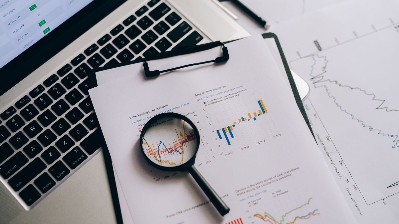 Lupa sobre relatórios e gráficos financeiros ao lado de um notebook, representando a comparação de taxas e prazos de CDBs.