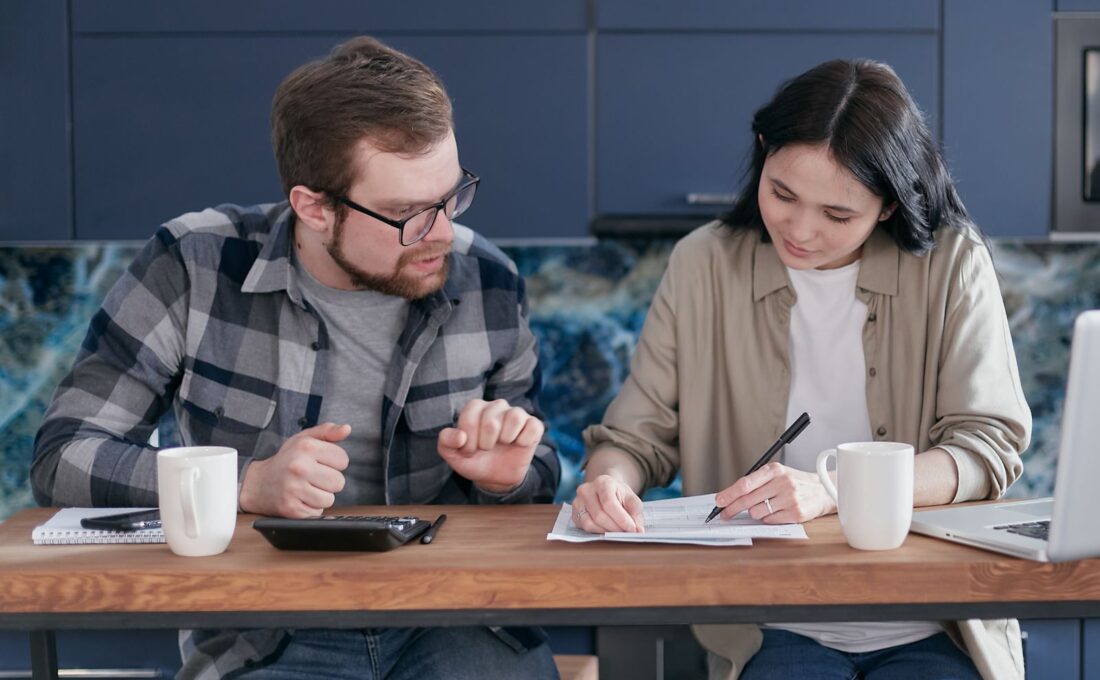 Duas pessoas analisam documentos financeiros com notebook e calculadora em uma mesa