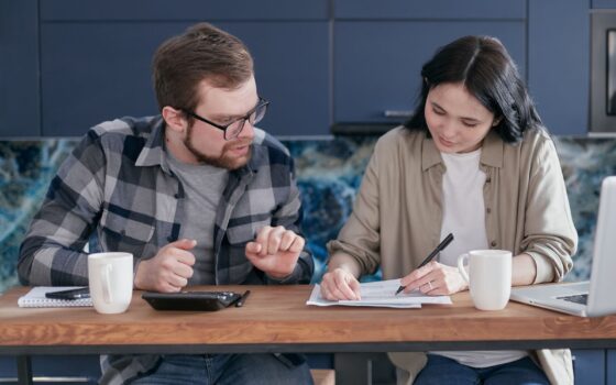 Duas pessoas analisam documentos financeiros com notebook e calculadora em uma mesa