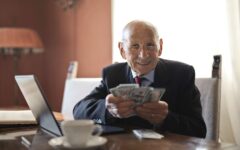 Homem idoso de óculos segura cédulas diante de um notebook em uma mesa