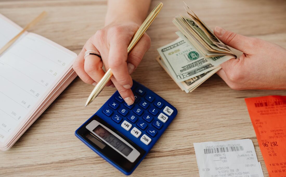 Pessoa anotando despesas ao lado de calculadora, cédulas e moedas em uma mesa