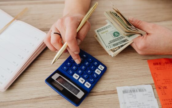 Pessoa anotando despesas ao lado de calculadora, cédulas e moedas em uma mesa