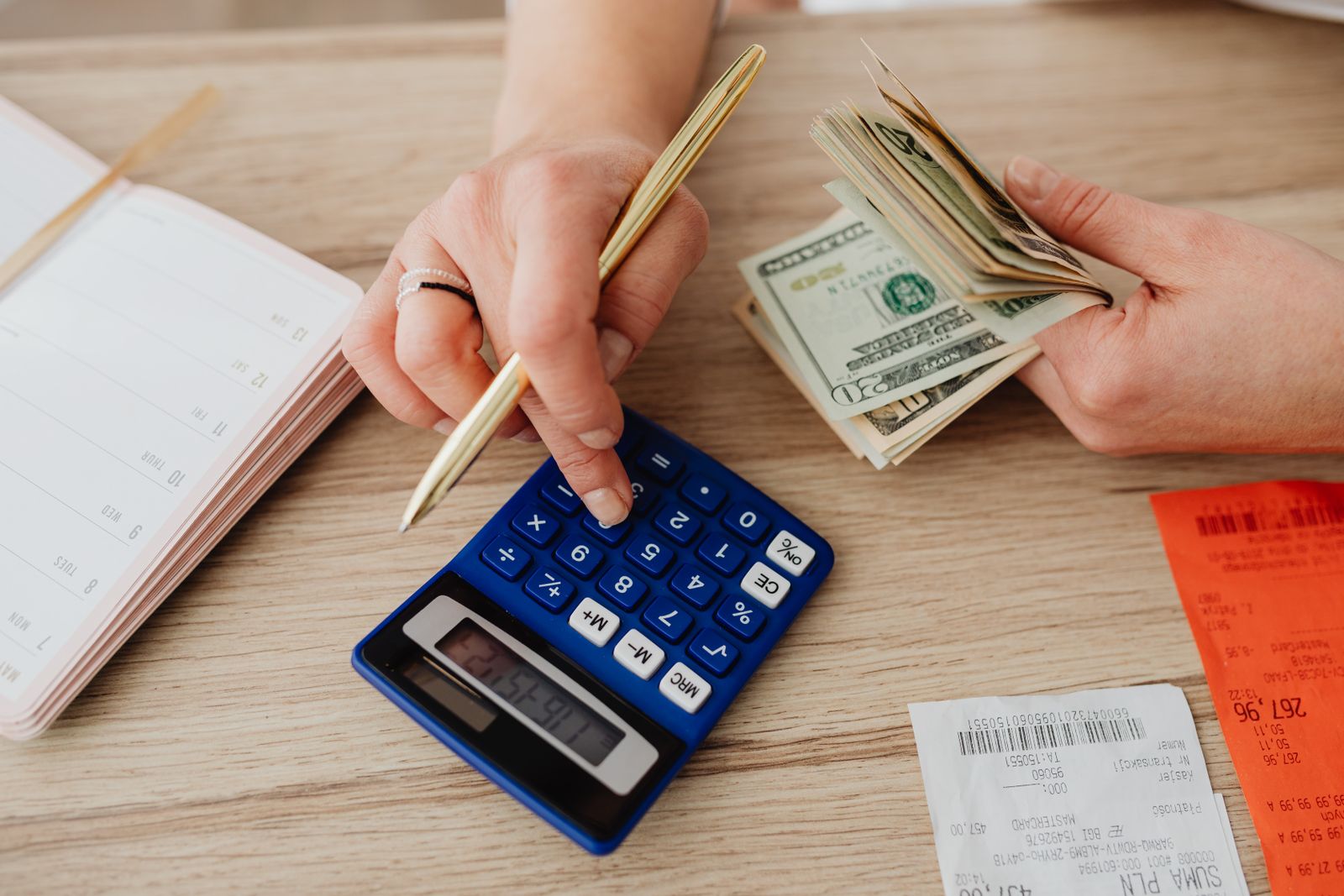 Pessoa anotando despesas ao lado de calculadora, cédulas e moedas em uma mesa