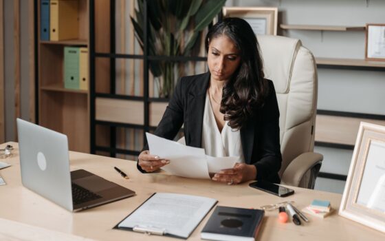 Mulher sentada à mesa de escritório analisa documentos impressos, imagem ilustrativa para a organização de informações trabalhistas