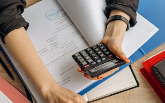 Pessoa usa uma calculadora ao lado de um notebook e documentos em uma mesa.