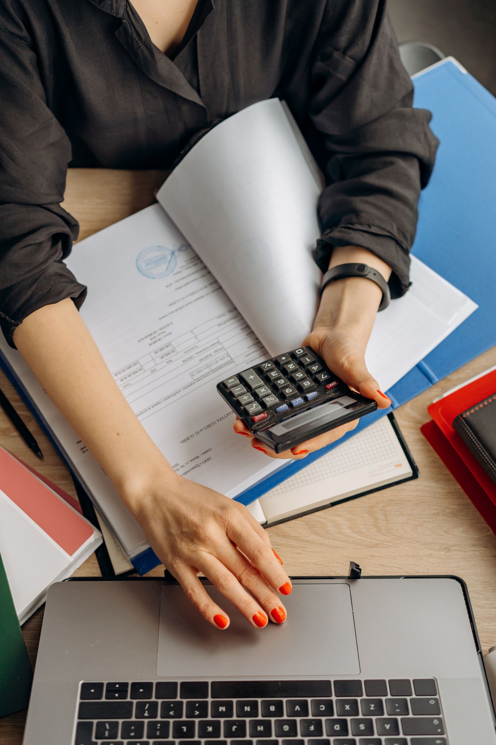 Pessoa usa uma calculadora ao lado de um notebook e documentos em uma mesa.