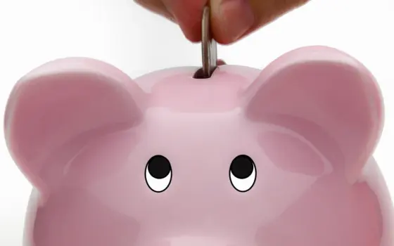 Pessoa colocando uma moeda em um cofrinho sobre uma mesa, imagem ilustrativa para guardar dinheiro e investir