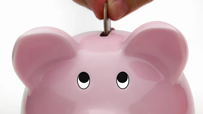 Pessoa colocando uma moeda em um cofrinho sobre uma mesa, imagem ilustrativa para guardar dinheiro e investir