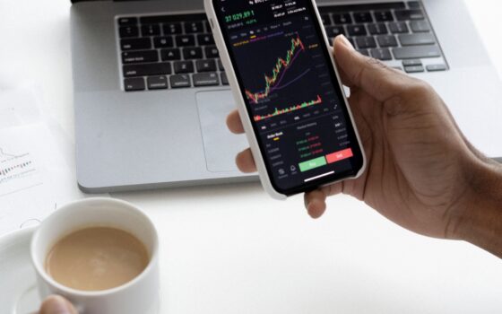 Pessoa analisa gráficos financeiros em um smartphone ao lado de um notebook