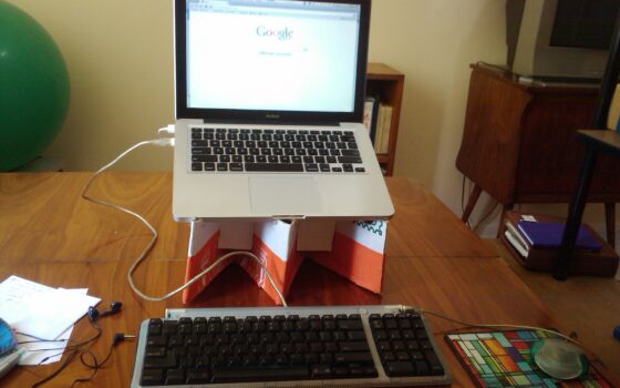 Mesa de trabalho em casa com laptop sobre suporte de papelão e caderno