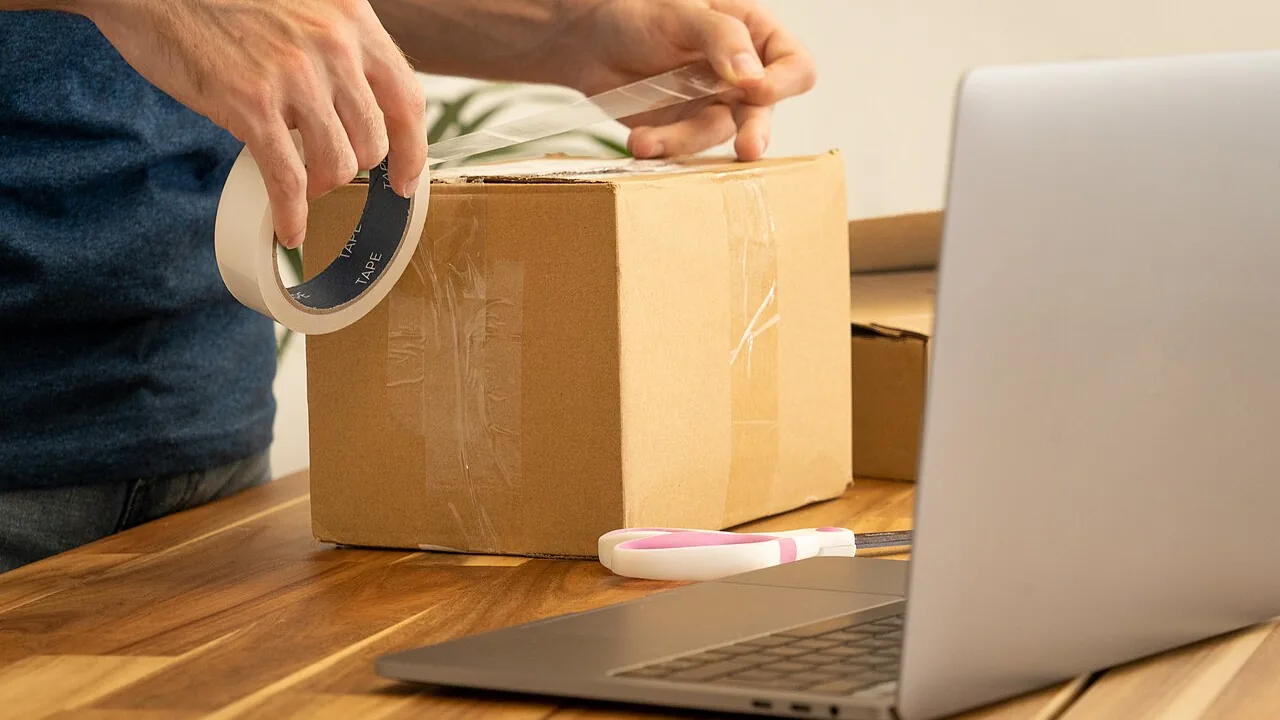 Pessoa fechando uma caixa de encomenda ao lado de um laptop
